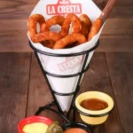 Professional food photography of La Cresta fried onion rings in a branded paper cone, served with three dipping sauces and a green chili pepper.