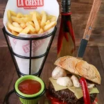 A la Cresta food photo session capturing a large sandwich with a knife, fries in a branded cone, and chili oil.