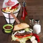 Food photography of a La Cresta sandwich, fries in a branded paper cone, chili oil, and seasonings on a wooden surface.