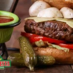 Food photography of a La Cresta burger sandwich, tomatoes, and mushrooms, served with pickles and a side of red sauce.